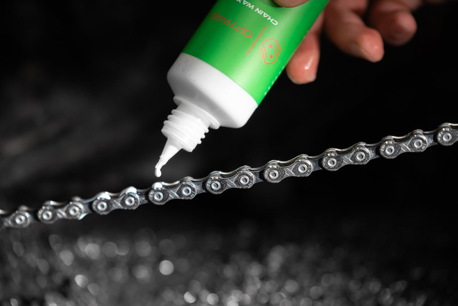 Eine Hand trägt vor einem dunklen, unscharfen Hintergrund Schmiermittel aus einer grünen Flasche auf eine silberne Fahrradkette auf.
