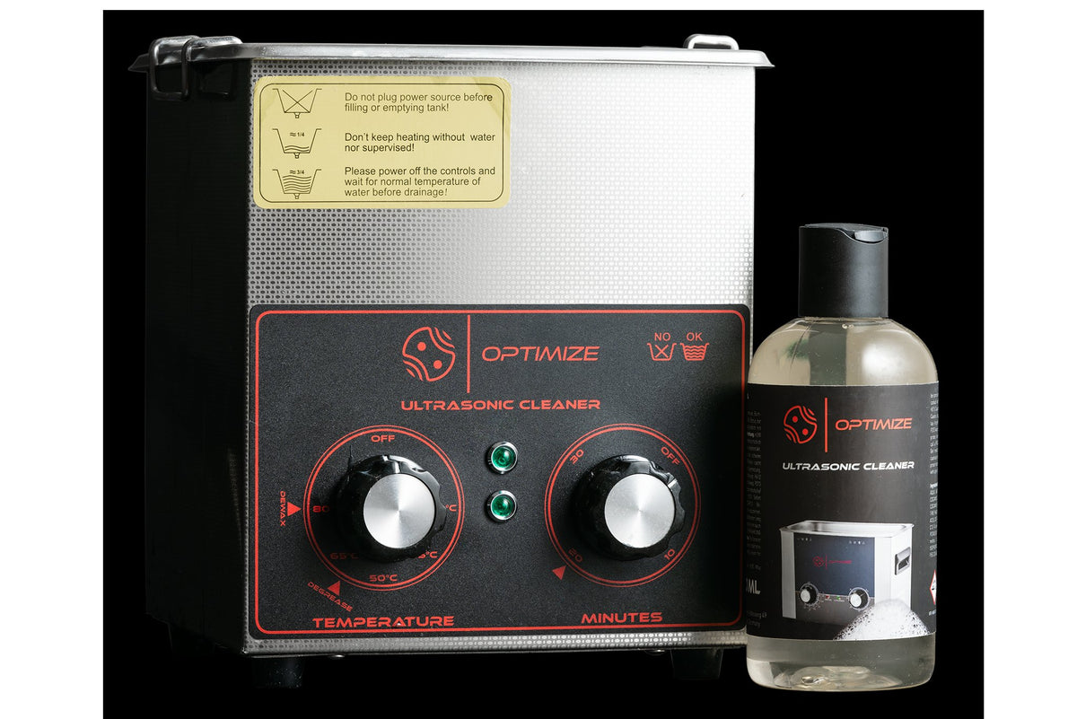 Ein Metall-Ultraschallreiniger mit zwei Drehknöpfen und der Aufschrift Optimize ist neben einer durchsichtigen Flasche mit Reinigungslösung mit einem passenden Etikett abgebildet, beide vor einem schwarzen Hintergrund.