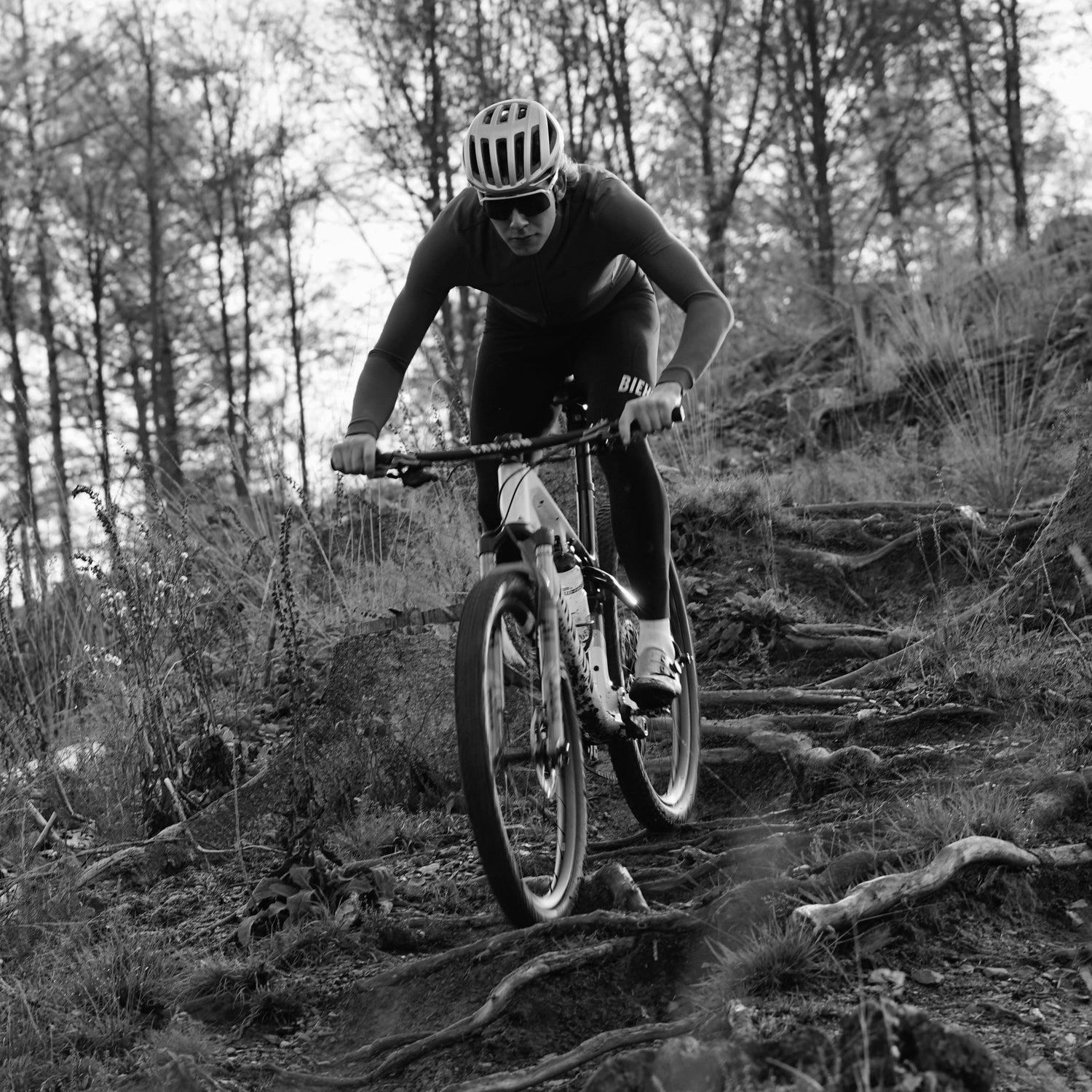 Eine Person mit Helm und Sonnenbrille fährt auf einem Mountainbike einen zerklüfteten, wurzelübersäten Waldweg hinunter. Das Bild ist in Schwarz-Weiß gehalten und fängt den Nervenkitzel der Fahrt ein, der durch die reibungslose Leistung des Kettenwachses auf der Kette noch verstärkt wird.