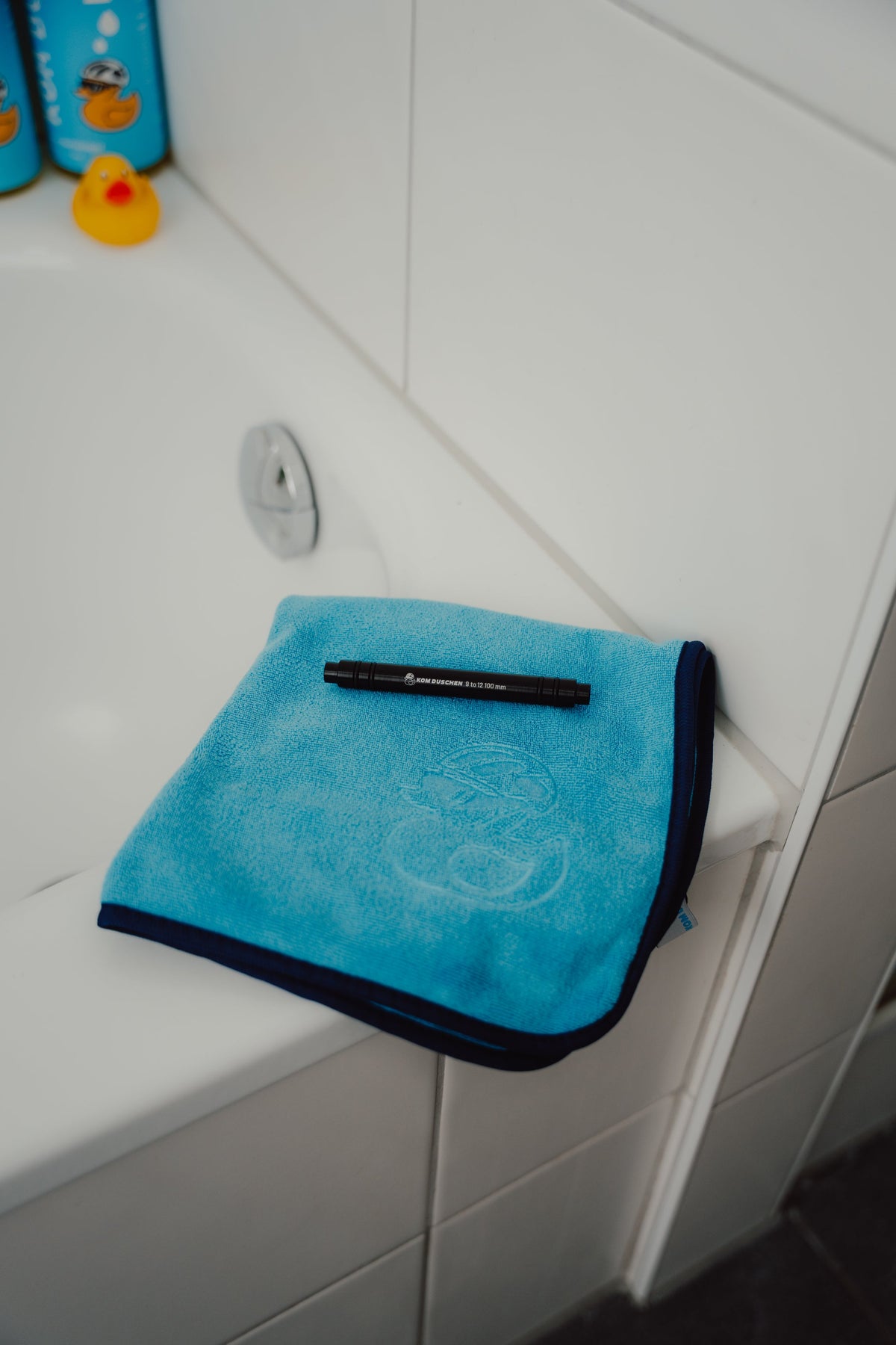 Ein hochwertiges Mikrofasertuch der KOM Duschen GmbH – Premium 800 g/m² mit dunkelblauem Rand – liegt am Rand einer weißen Badewanne, darauf liegt ein schwarzer Stift.