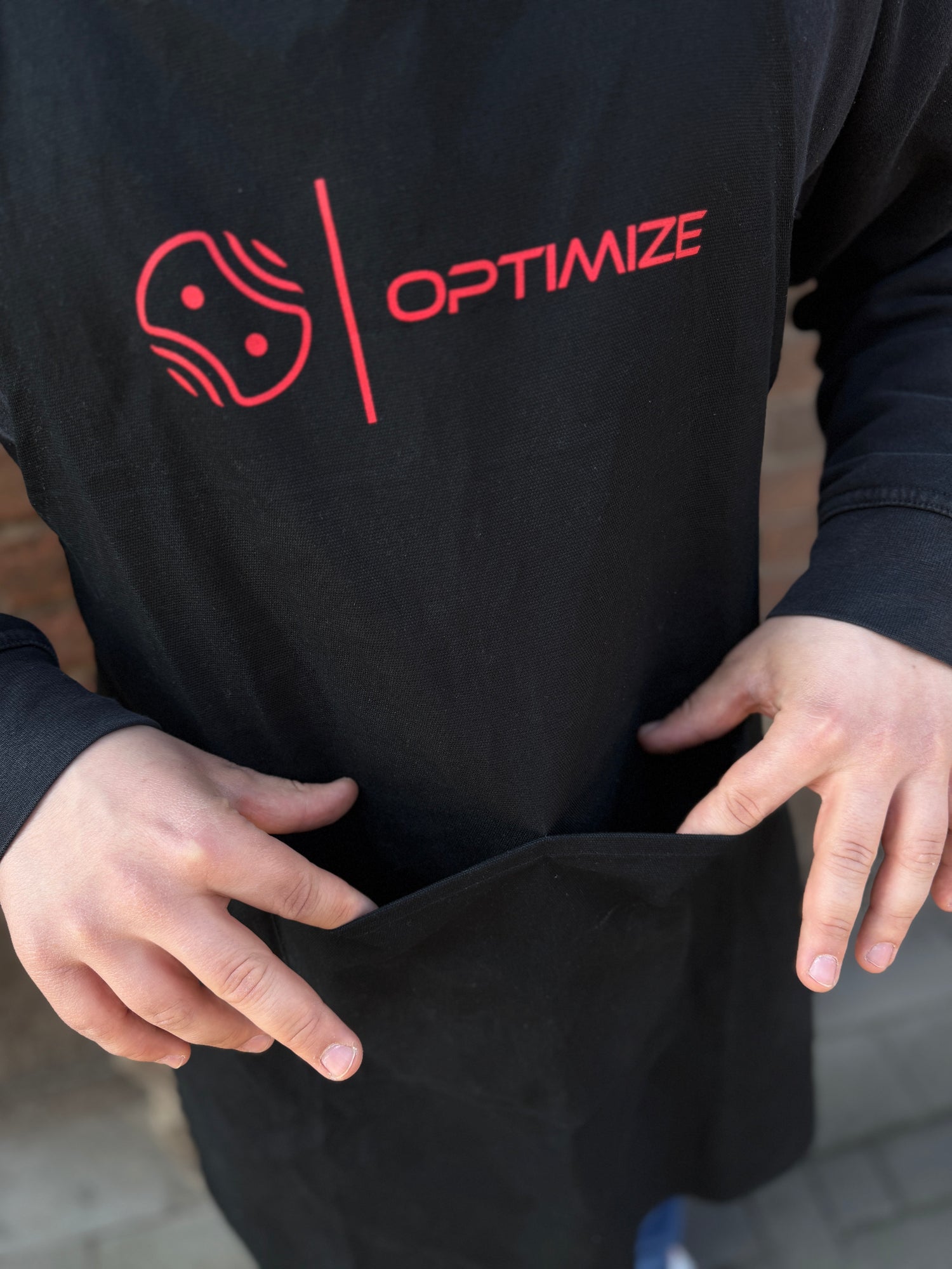 Eine Person trägt die schwarze Optimize Heißwachsschürze von Mein Shop mit rotem OPTIMIZE-Logo und -Symbol, während sie die Vordertasche offen hält; nur ihre Hände und ihr Oberkörper sind sichtbar.
