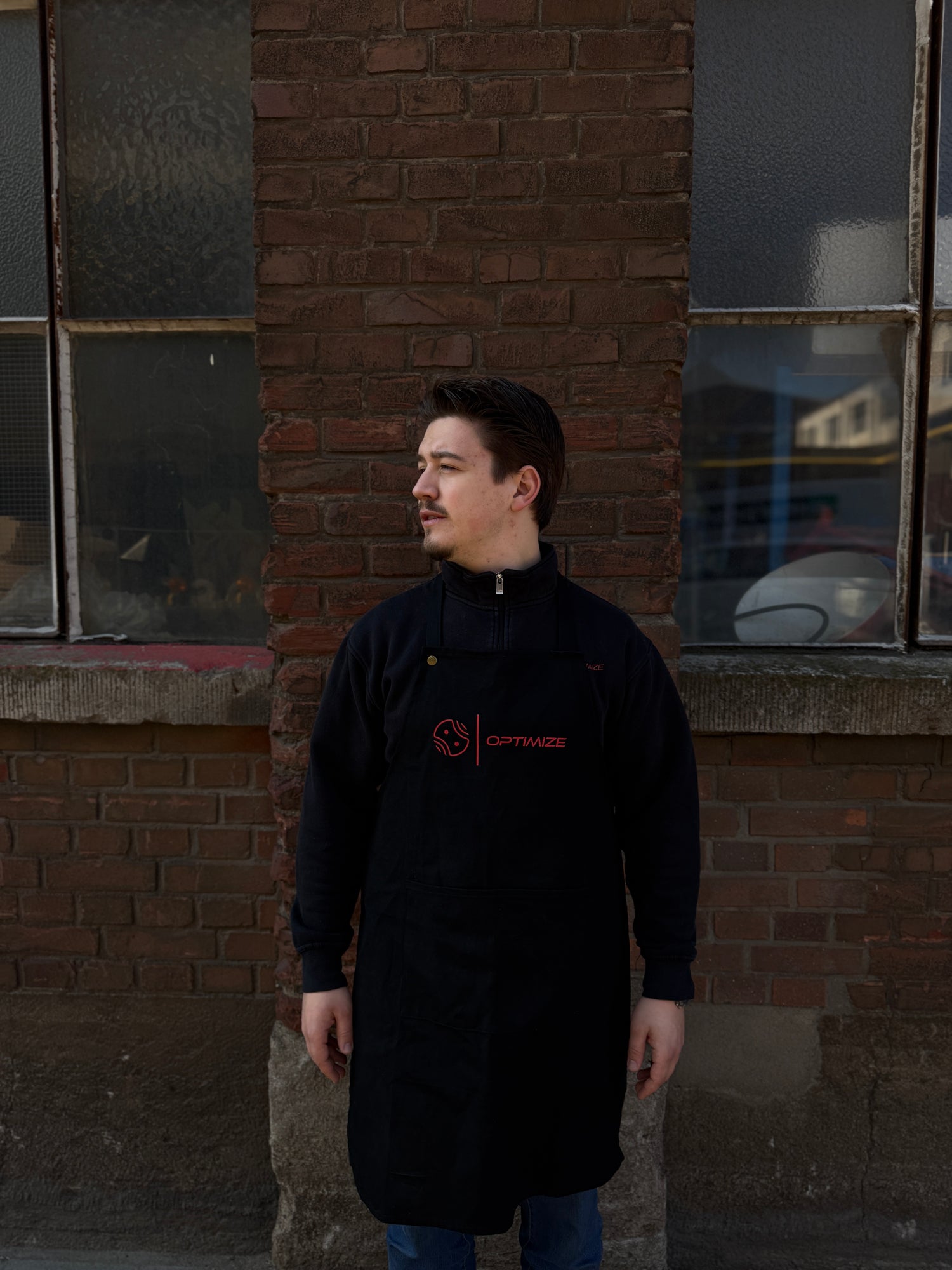 Ein Mann mit kurzen dunklen Haaren, der eine schwarze Mein Shop Optimize Kochschürze und eine Jacke trägt, steht an einer Backsteinmauer mit zwei großen Fenstern und blickt nach links, die Hände an den Seiten.