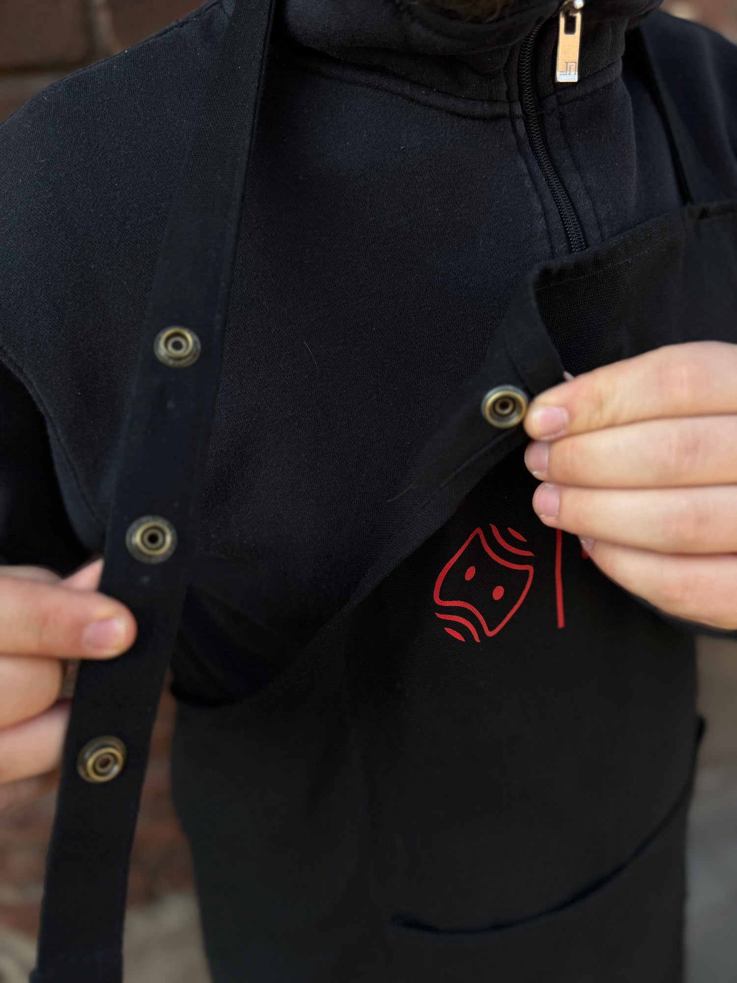Eine Person, die eine schwarze Jacke mit Reißverschluss trägt, schließt Metalldruckknöpfe an der Mein Shop Optimize Heißwachsschürze, einer schwarzen Schürze mit einem roten, abstrakten Gesichtsmuster auf der Brust, während sie die Träger festhält.