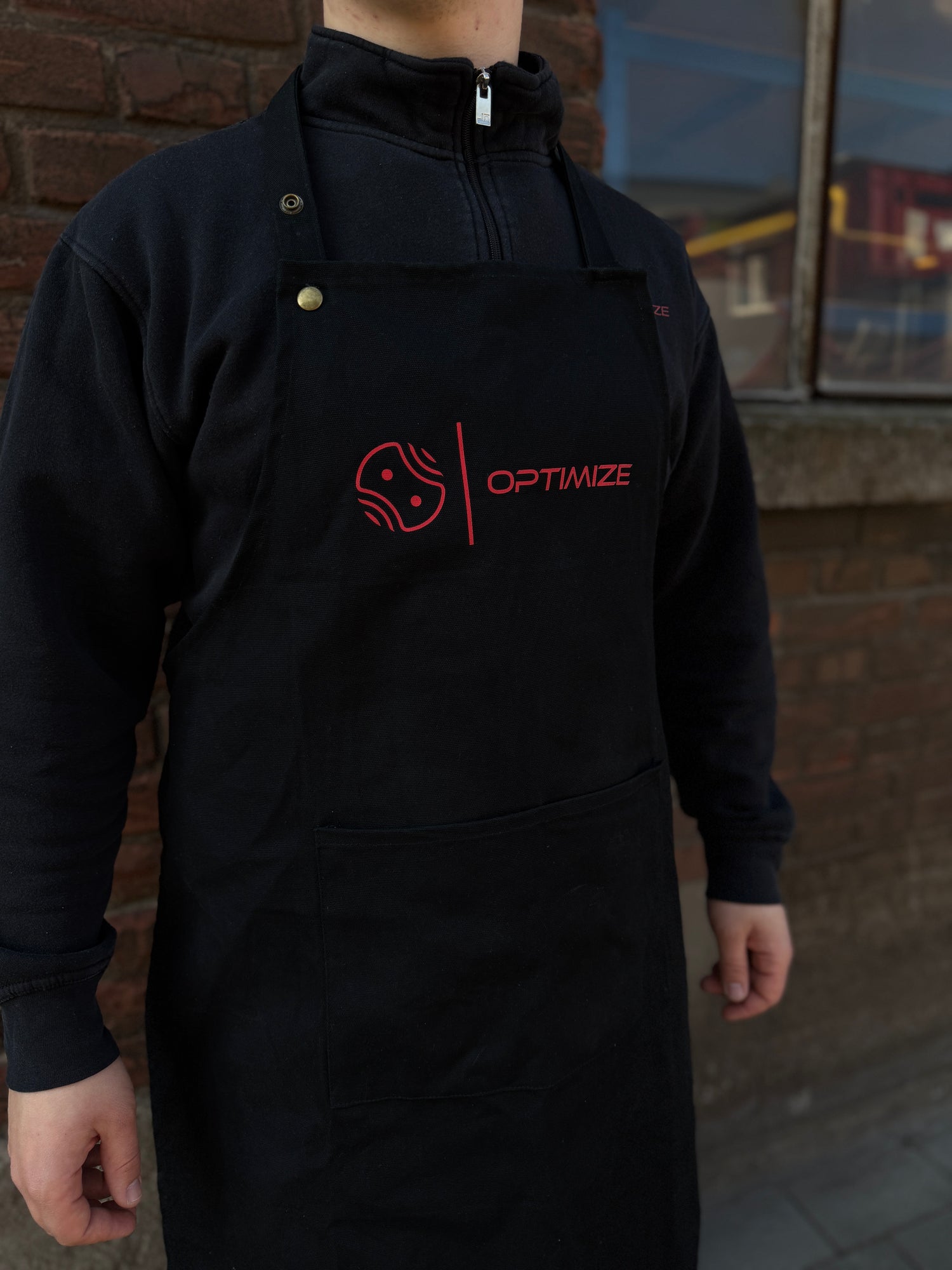 Eine Person trägt die schwarze Optimize Heißwachsschürze von Mein Shop mit rotem OPTIMIZE-Logo über einem schwarzen Pullover mit Reißverschluss, während sie draußen vor einer Backsteinmauer und einem Fenster steht.