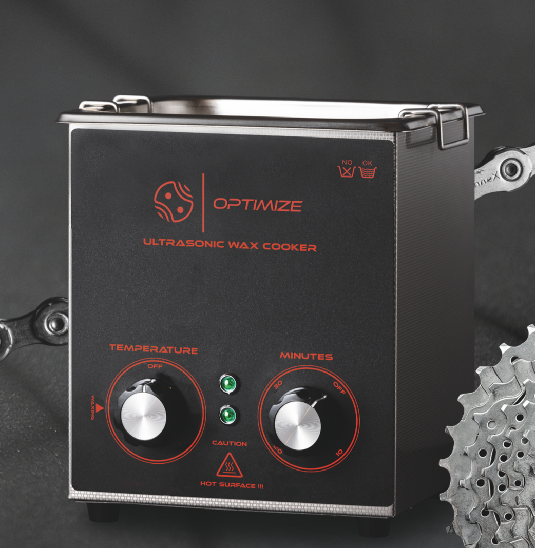 Ein schwarzer Ultraschall-Wachskocher mit OPTIMIZE-Branding, zwei Drehreglern für Temperatur und Minuten, grünen Kontrollleuchten und einer Fahrradkette und -kassette aus Metall - ideal für Kettenwachs-Anwendungen - ist daneben teilweise sichtbar.