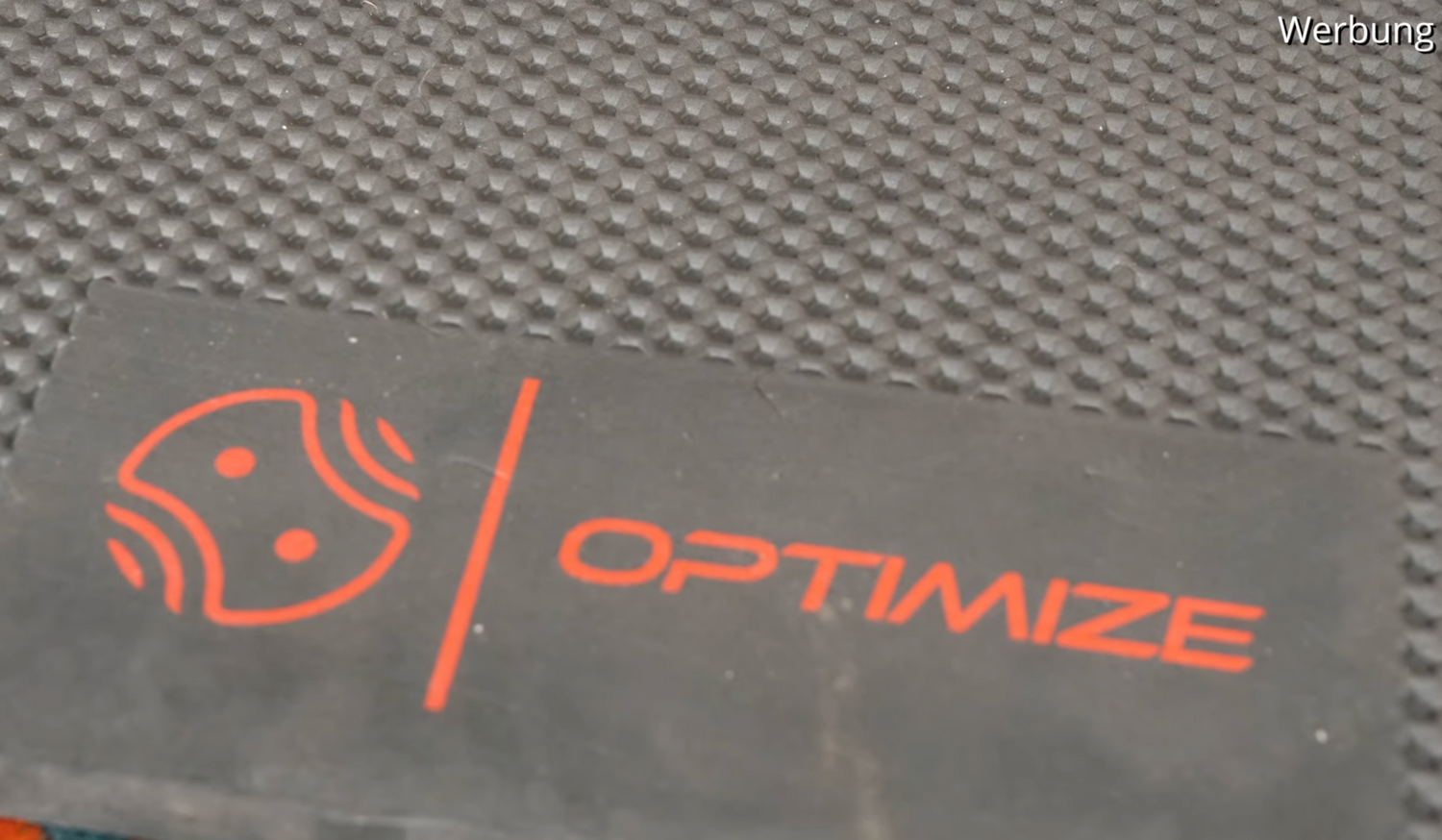 Eine Nahaufnahme der Indoor Matte der Optimize GmbH zeigt eine strukturierte schwarze Oberfläche mit einem rechteckigen schwarzen Etikett, das ein rotes abstraktes Logo und das fette rote Wort