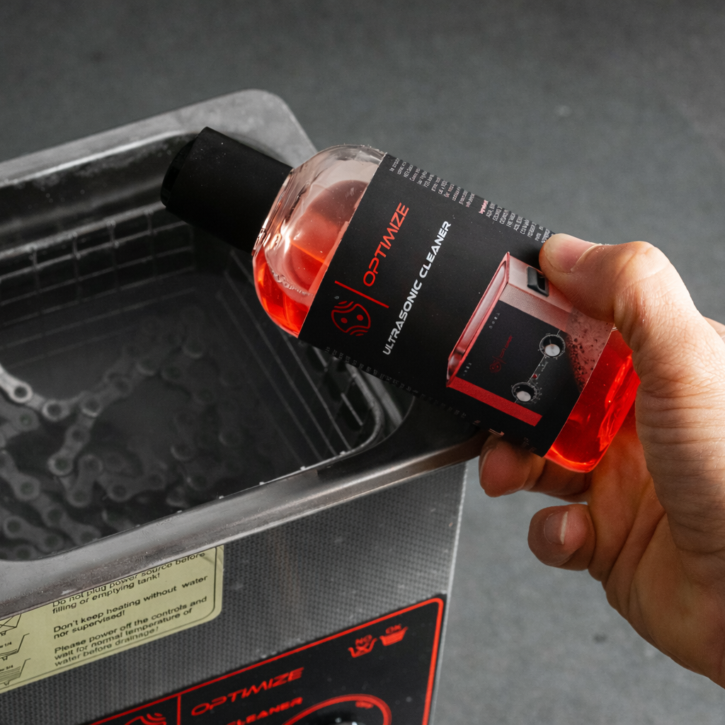 Eine Hand gießt rote „Optimize Ultrasonic Cleaner“-Flüssigkeit aus einer Flasche in ein mit Wasser gefülltes „Optimize“-Ultraschallbad – ideal für eine gründliche Kettenreinigung und ein effektives Entfetten der Kette.