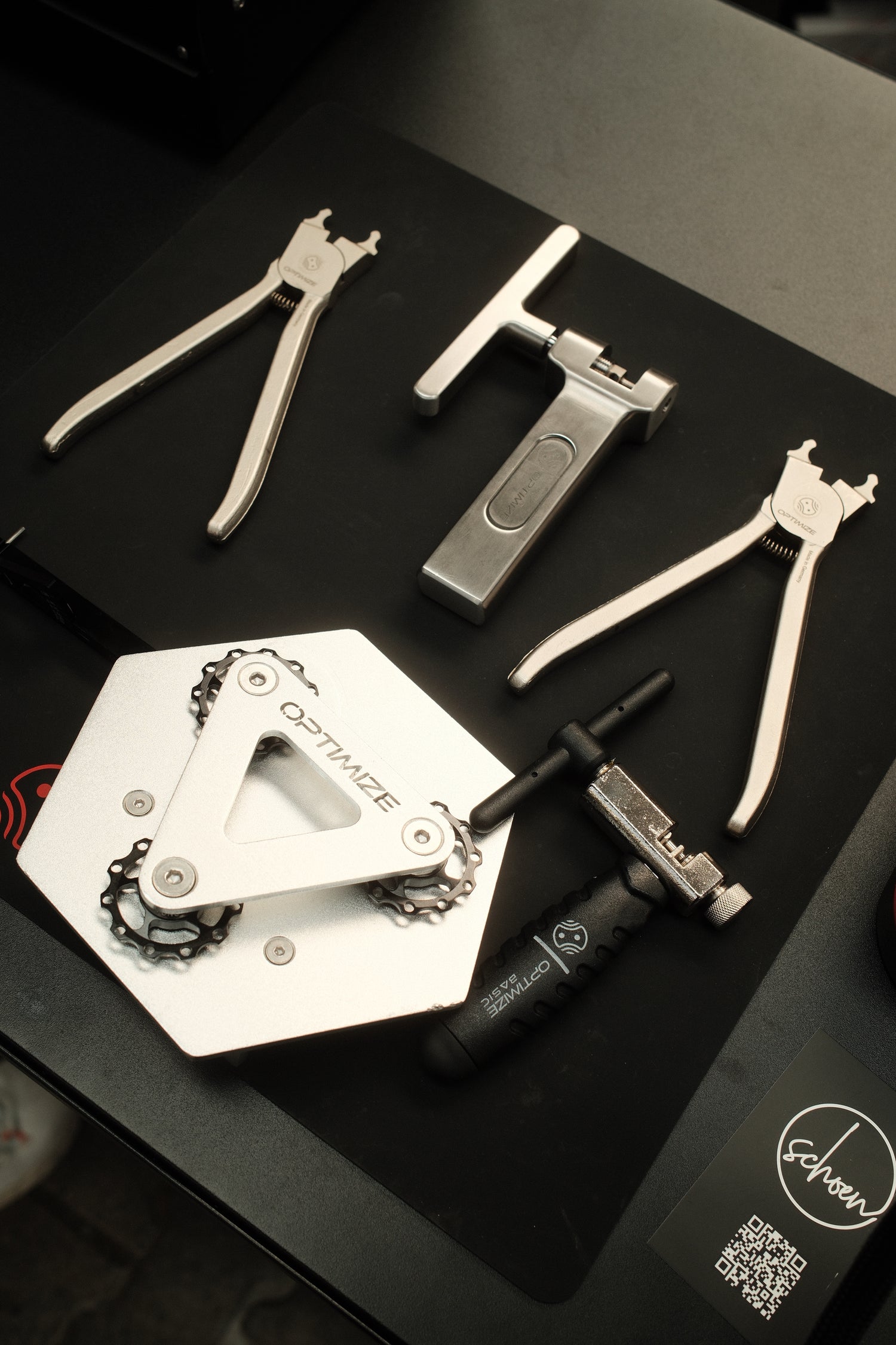 Ein Satz Fahrradreparaturwerkzeuge der Optimize GmbH, darunter der Optimize Kettenbrecher mechanisch, eine Zange und ein Kettenhalter, angeordnet auf einer schwarzen Fläche mit einem Eshon-Aufkleber und einem QR-Code daneben.