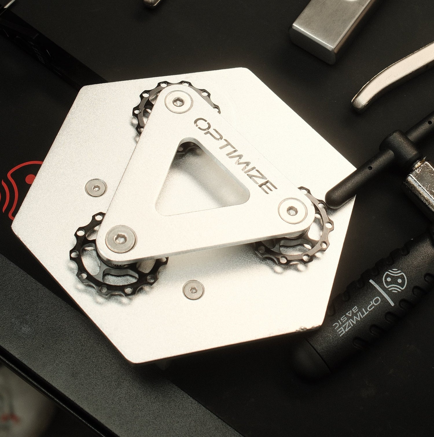 Ein sechseckiger metallischer Kettenbrecher mechanisch von Optimize GmbH, mit drei kleinen Zahnrädern und der Gravur OPTIMIZE
