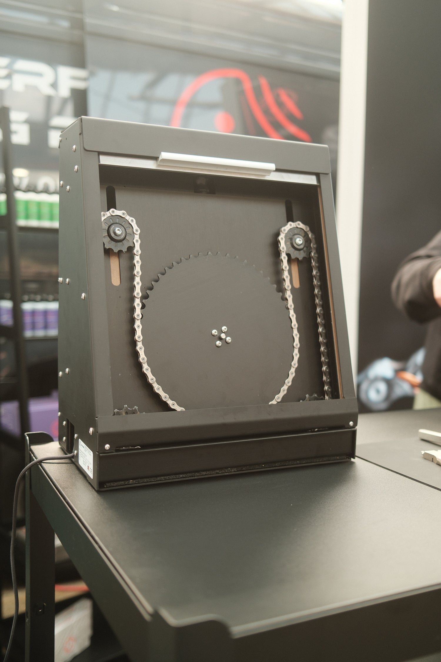 Der Optimize Kettenbrecher elektrisch der Optimize GmbH, ein schwarzes mechanisches Gerät mit einem großen Kreissägeblatt und sichtbaren Kettenmechanismen, steht auf einem Tisch mit Regalen mit bunten Artikeln, die im Hintergrund verschwimmen.