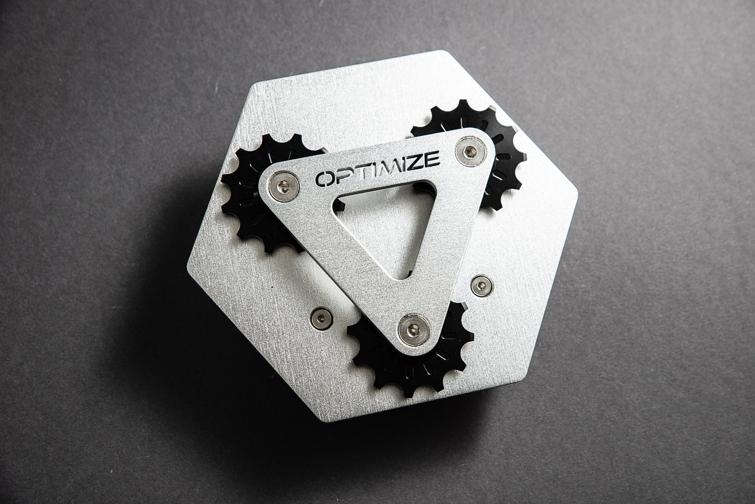 Der „Kettenbrecher mechanisch“ von Optimize GmbH ist ein metallisches, sechseckiges Objekt mit drei schwarzen Zahnrädern, einem dreieckigen Metallteil mit der Gravur „OPTIMIZE“ sowie Designelementen, die von einem mechanischen Kettenbrecher inspiriert sind, vor einem dunkelgrauen Hintergrund.