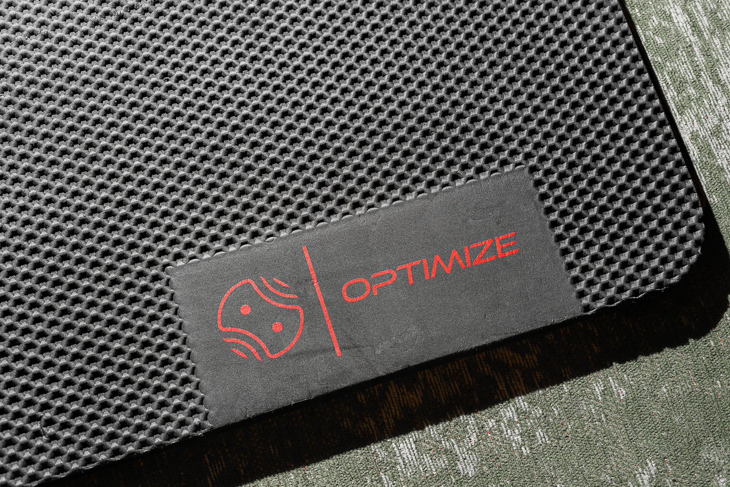 Eine Nahaufnahme eines strukturierten schwarzen „Indoor Matte“-Produkts der Optimize GmbH, auf dem ein Etikett mit einem roten Logo und dem Schriftzug „OPTIMIZE“ in Rot auf einem grünen Untergrund zu sehen ist.