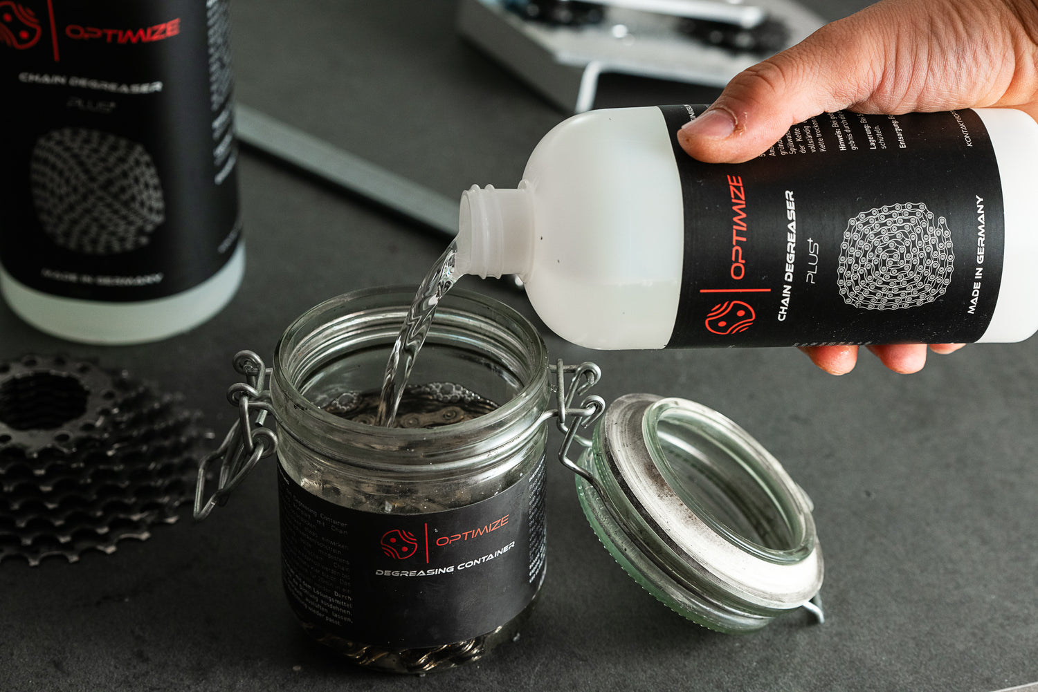 Eine Hand gießt „Chain Degreaser Plus“ aus einer weißen Flasche mit der Aufschrift „Optimize“ in den „Optimize“-Entfettungsbehälter, in dem sich Metallteile eines Fahrrads befinden; eine weitere Flasche und zusätzliche Teile sind auf einer grauen Fläche in der Nähe zu sehen, die sich ideal für die Lagerung von Lösungsmitteln eignet (Lösungsmittel Lagerung).