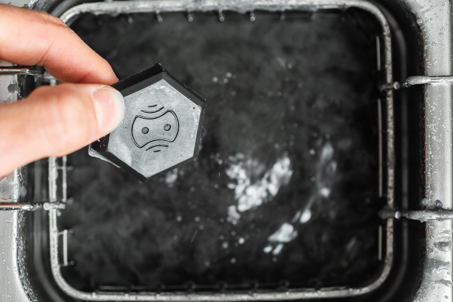 Eine Hand hält eine schwarze sechseckige Marke mit einem traurigen Gesichtssymbol über einem mit Wasser gefüllten Waschbecken, das an die Verwendung von Optimize Entfettungs-/Entfettungsadditiv von Optimize während der Reinigungs- oder Wartungsroutinen erinnert.