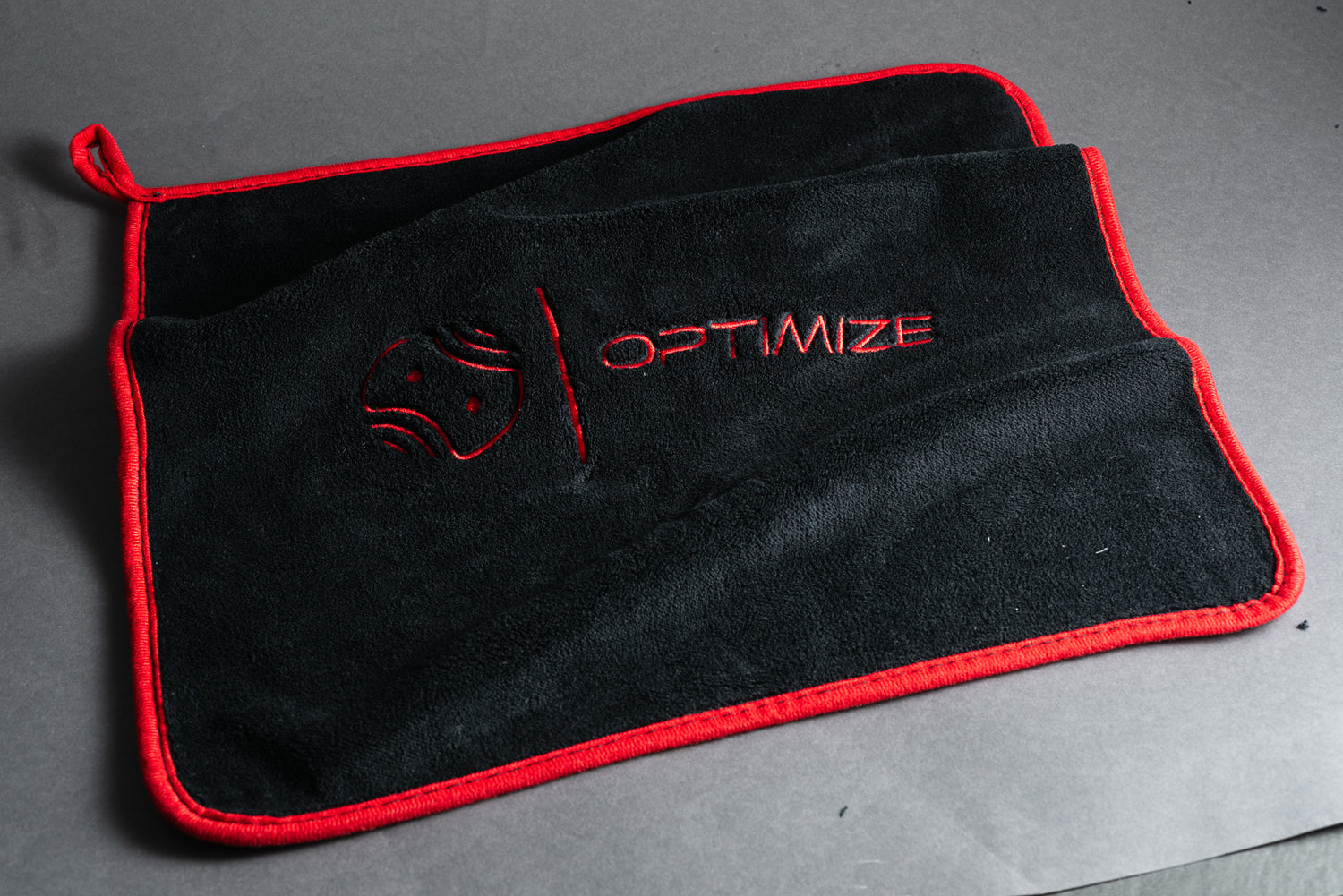 Das schwarze Optimize Premium Mikrofasertuch von Optimize GmbH mit rotem Rand und eingewebtem Logo sowie