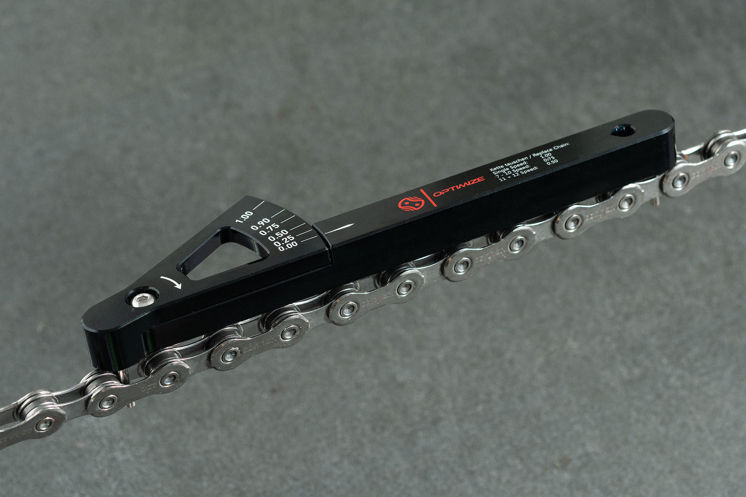 Die Optimize Kettenverschleißlehre der Optimize GmbH ist in schwarzer Farbe auf einer silbernen Shimano-, KMC- oder Connex-Fahrradkette über einer grauen Oberfläche abgebildet und zeigt Kettendehnungsmarkierungen zur präzisen Messung an.
