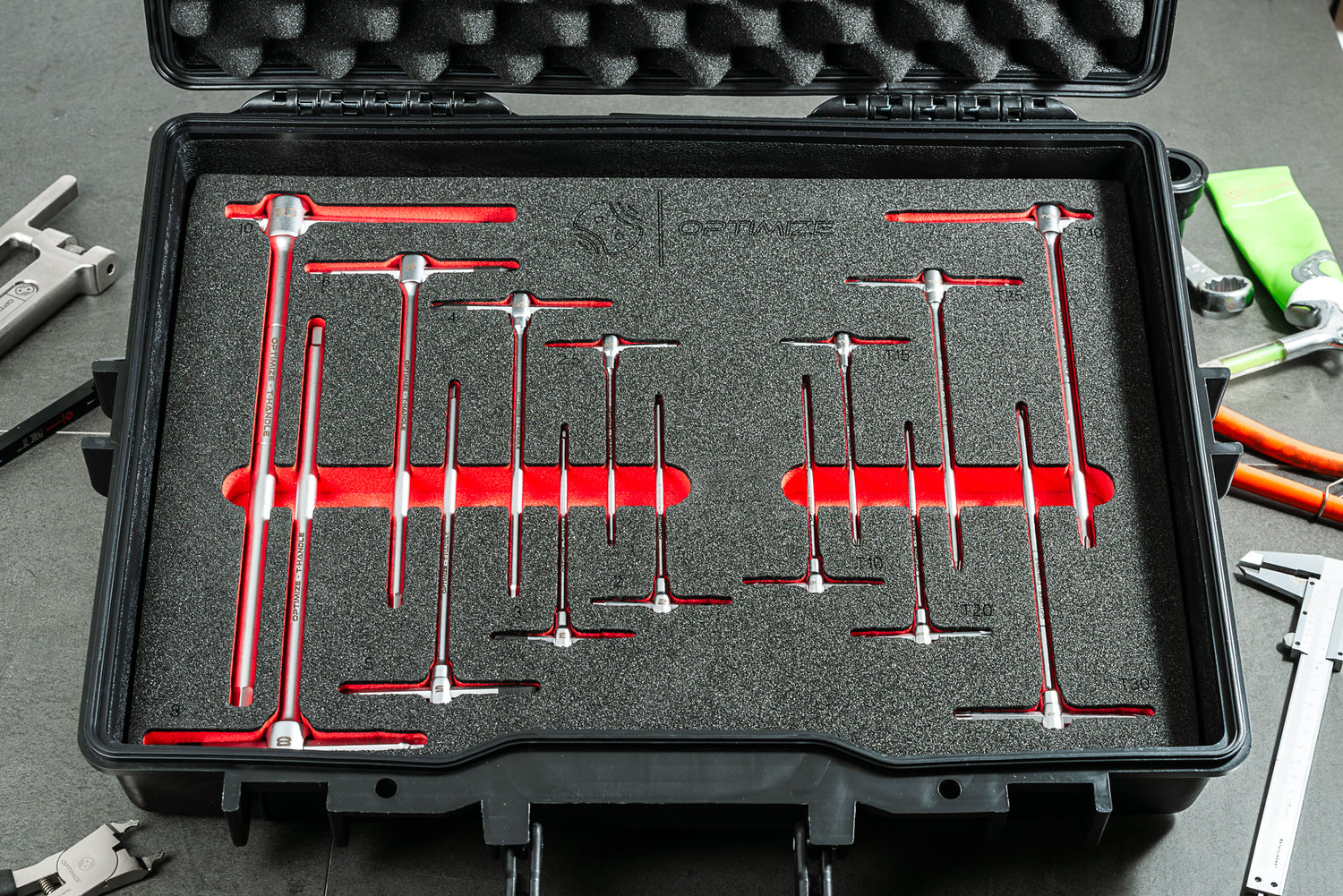 Der Optimize Werkzeugkoffer von Optimize GmbH verfügt über Schaumstoffeinlagen, die verschiedene T-Griff-Inbusschlüssel aufnehmen, ideal als Bike-Werkzeug. Der schlichte schwarze Koffer liegt offen auf einer Werkbank zwischen anderen Werkzeugen und einem grünen Tuch.