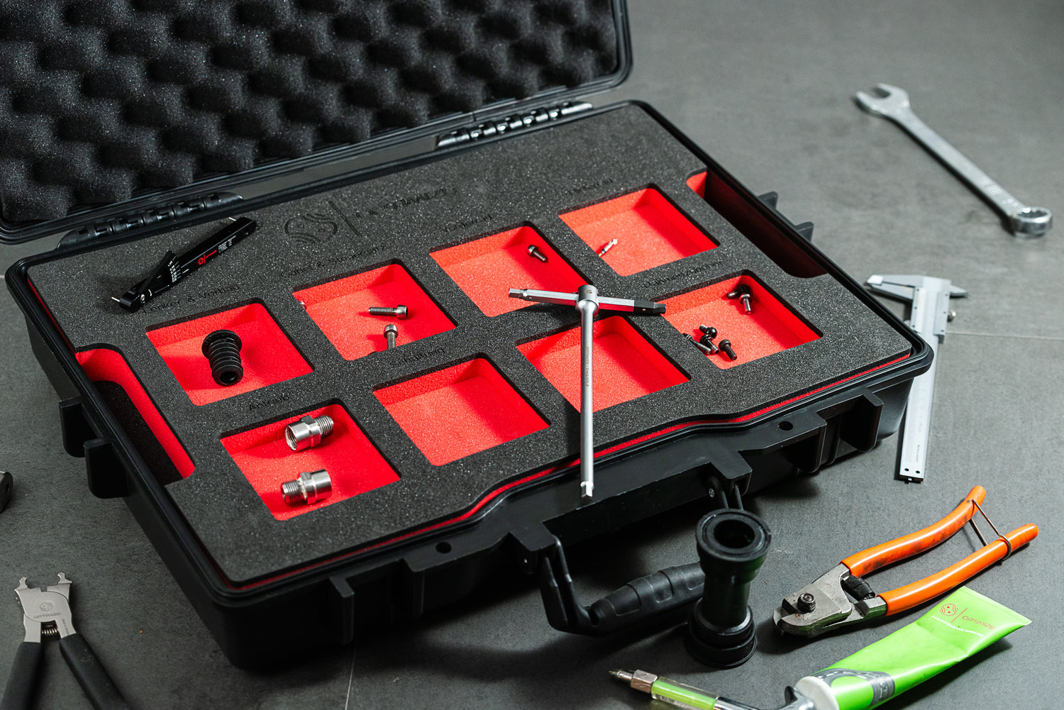 Ein Optimize Werkzeugkoffer der Optimize GmbH, mit Schaumstoffeinlagen, nimmt kleine Metallwerkzeuge wie Schraubenschlüssel und Beschläge auf. Außerhalb des schwarzen Koffers befinden sich Zangen, ein Schraubenschlüssel, ein grünes Werkzeug und andere sortierte Werkzeuge auf einer grauen Fläche.