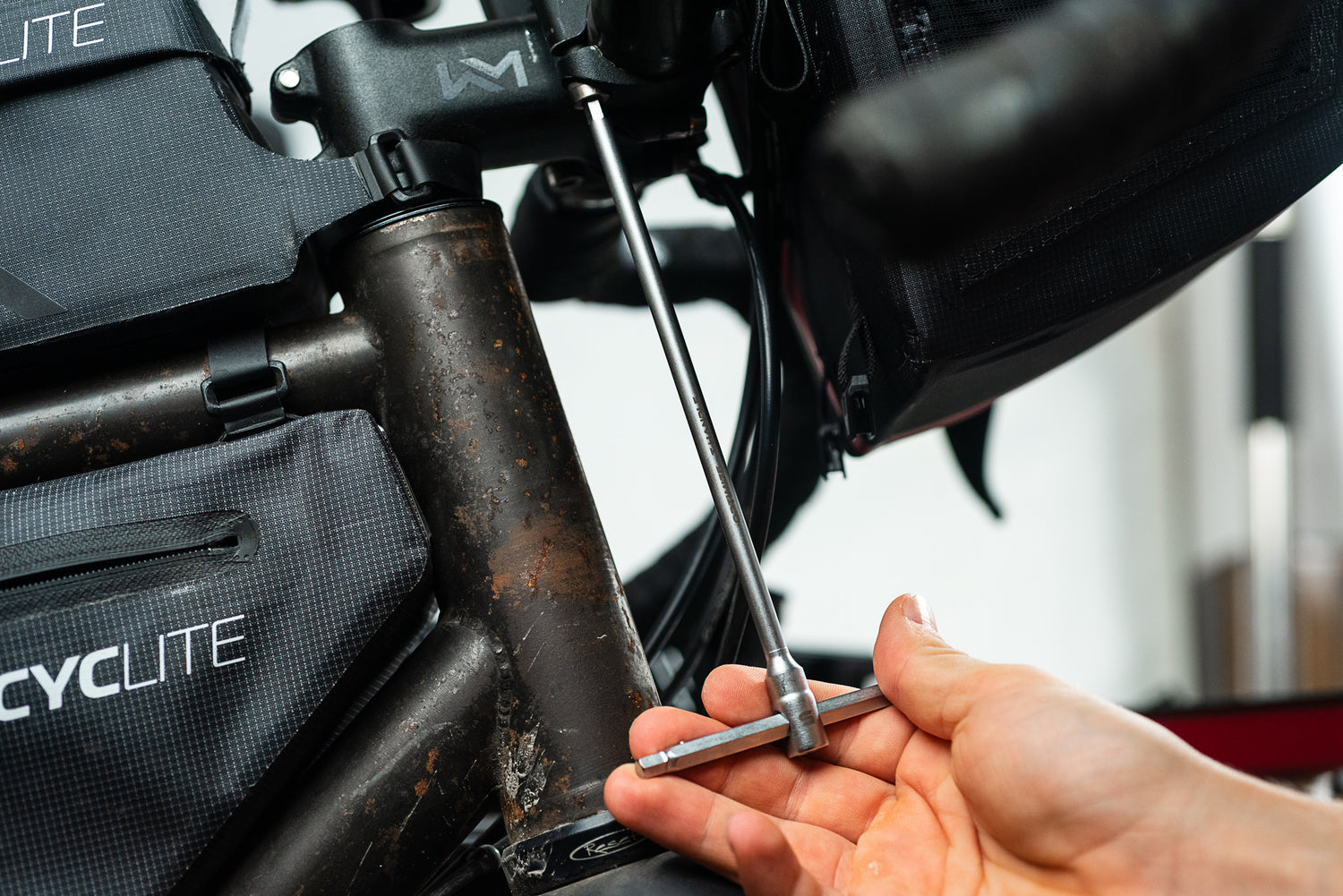 Eine Hand benutzt einen T-Griff-Sechskantschlüssel aus dem Optimize Werkzeugkoffer der Optimize GmbH, um eine Schraube an einem verwitterten Fahrradrahmen, der mit schwarzen CYC LITE Packs ausgestattet ist, einzustellen.