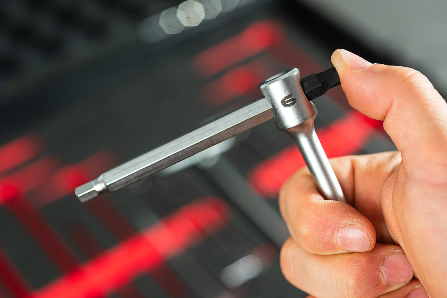 Eine Hand hält einen silbernen L-förmigen Innensechskantschlüssel mit schwarzem Bit aus dem Optimize Werkzeugkoffer der Optimize GmbH, ideal für jedes Ordnungssystem, vor einem unscharfen roten und schwarzen Hintergrund.