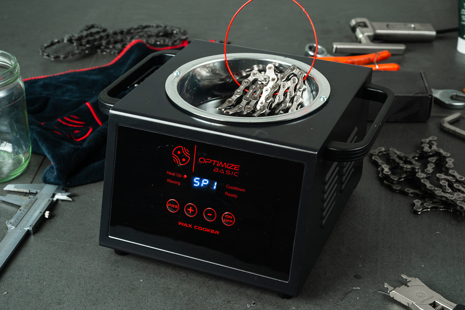 Eine Fahrradkette wird in einen Optimize Automatischen Wachskocher der Optimize GmbH getaucht und mit Fahrradwerkzeug und losen Kettengliedern auf eine Werkbank gelegt. Das digitale Display zeigt den Heizstatus für effizientes Wachsen der Kette an.