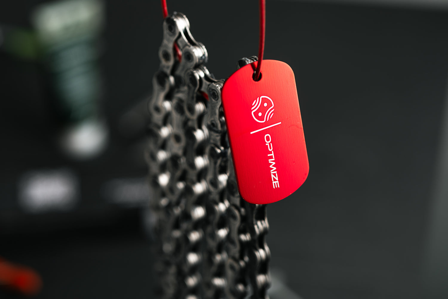 Eine Nahaufnahme zeigt eine Fahrradkette mit einem Optimize Chain Tag der Optimize GmbH, die an einem dünnen roten Draht hängt. Der Hintergrund ist unscharf und dunkel und lenkt die Aufmerksamkeit auf die Kette und den Anhänger.