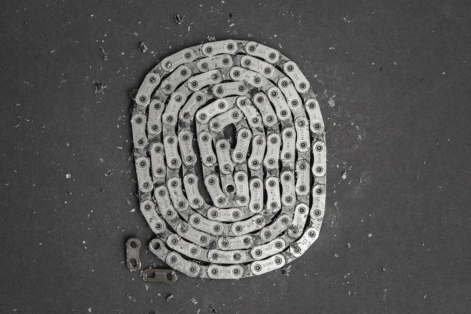 Eine vorgewachste SRAM Ketten Fahrradkette von Optimize GmbH ist in einer Spirale auf einer strukturierten dunklen Oberfläche aufgewickelt, mit mehreren getrennten Gliedern in der Nähe.