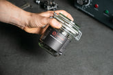 Eine Hand hält einen 400-ml-Optimize-Entfettungsbehälter mit einem schwarzen Optimize-Etikett vor einer grauen Fläche und unscharfen Maschinen im Hintergrund - perfekt für die Lagerung von Kettenentfetter Plus oder Lösungsmitteln.