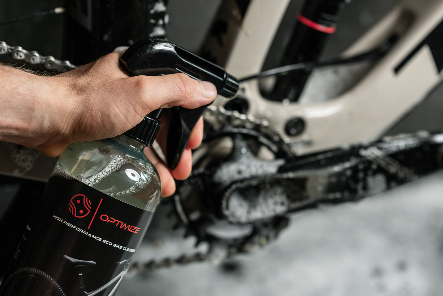 Eine Hand sprüht Optimize Fahrradreiniger - Wash and Wax von Optimize auf den Antriebsstrang und die Schaltung eines Fahrrads, mit Seife und Wasser auf die Komponenten - ein wesentlicher Schritt bei der gründlichen Fahrradpflege.