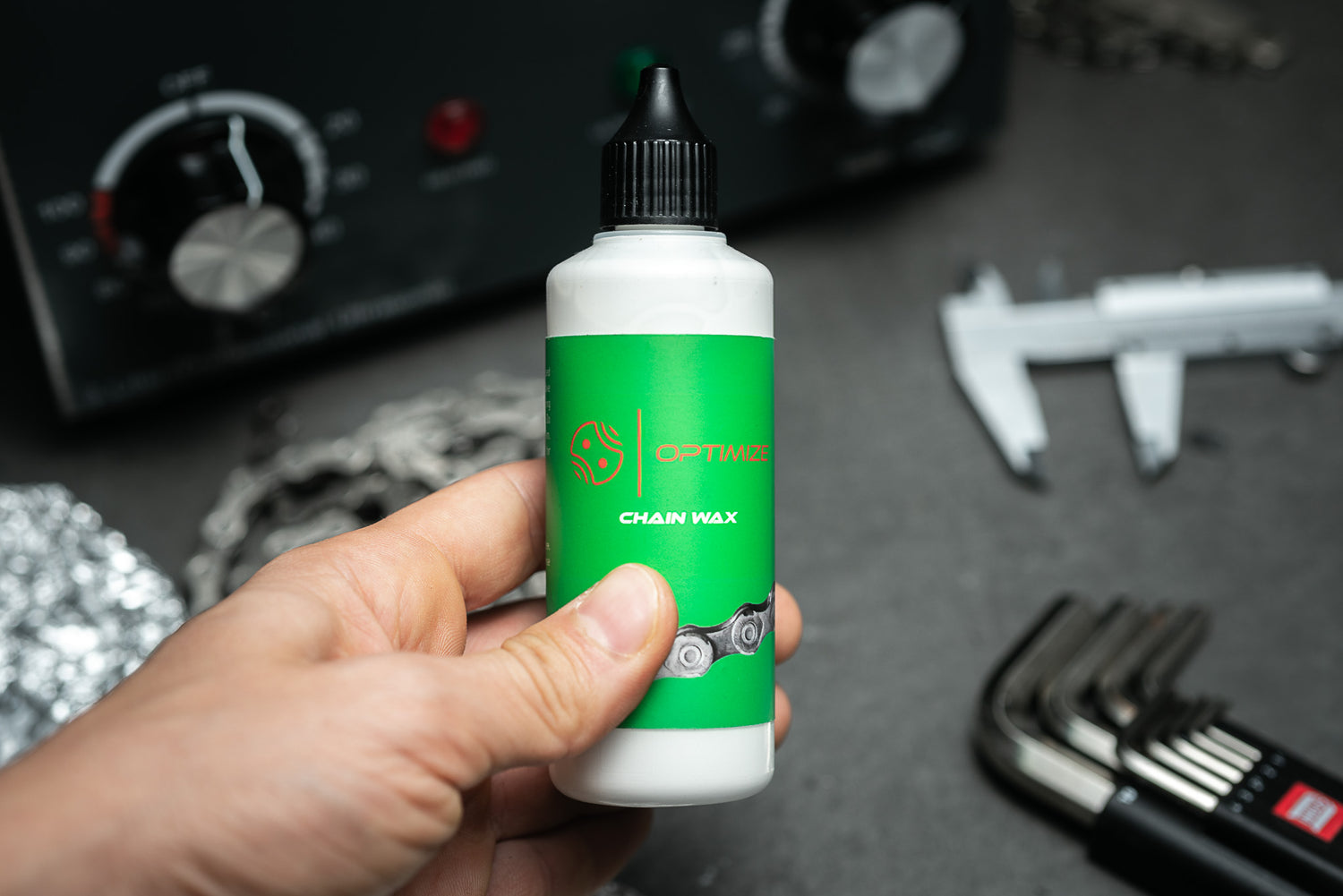 Eine Hand hält eine Flasche Mein Shop Optimize Kettenwachs mit einem grünen Etikett. Im Hintergrund sind Werkzeuge wie ein Messschieber, Inbusschlüssel, eine Fahrradkette und ein Bedienfeld zu sehen. Ideal für die zuverlässige Kettenschmierung.