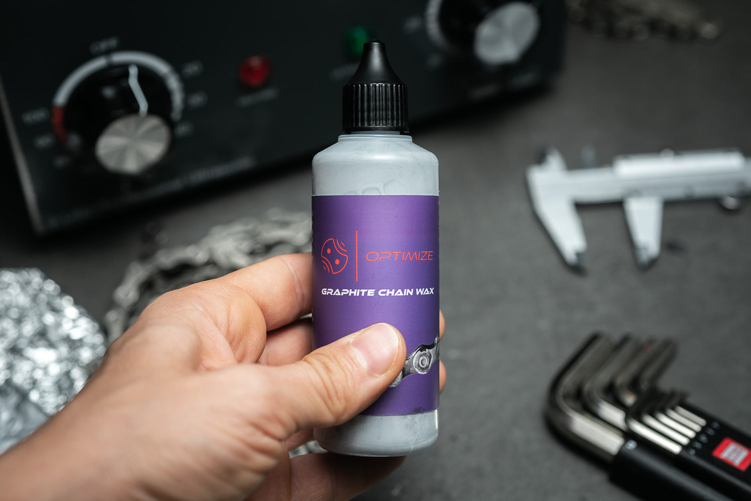 Eine Hand hält eine Flasche Mein Shop Optimize Graphit-Kettenwachs vor einem Hintergrund von Werkzeugen wie Messschiebern und Inbusschlüsseln, perfekt für die effiziente Fahrradkettenschmierung.