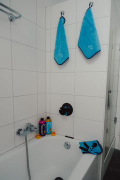 Zwei Mikrofasertücher der Marke KOM Duschen GmbH – Premium 800 g/m² – hängen an Haken über einer Badewanne, in der zwei Flaschen Duschgel und zwei Gummienten liegen.