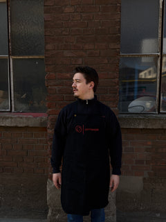 Ein Mann mit kurzen dunklen Haaren, der eine schwarze Mein Shop Optimize Kochschürze und eine Jacke trägt, steht an einer Backsteinmauer mit zwei großen Fenstern und blickt nach links, die Hände an den Seiten.