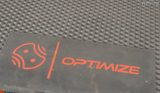 Eine Nahaufnahme der Indoor Matte der Optimize GmbH zeigt eine strukturierte schwarze Oberfläche mit einem rechteckigen schwarzen Etikett, das ein rotes abstraktes Logo und das fette rote Wort "OPTIMIZE" enthält. Oben rechts steht in kleiner Schrift "Werbung". Premium Dämpfung wird empfohlen.