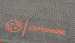 Eine Nahaufnahme der Indoor Matte der Optimize GmbH zeigt eine strukturierte schwarze Oberfläche mit einem rechteckigen schwarzen Etikett, das ein rotes abstraktes Logo und das fette rote Wort "OPTIMIZE" enthält. Oben rechts steht in kleiner Schrift "Werbung". Premium Dämpfung wird empfohlen.