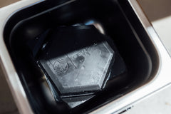 Ein schwarzes sechseckiges Objekt mit eingraviertem "OPTIMAX" und Logo ruht im Inneren des Optimize Ultraschall Wachskochers, ideal von Optimize zum effizienten Schmelzen von Heißwachs oder Kettenwachs.