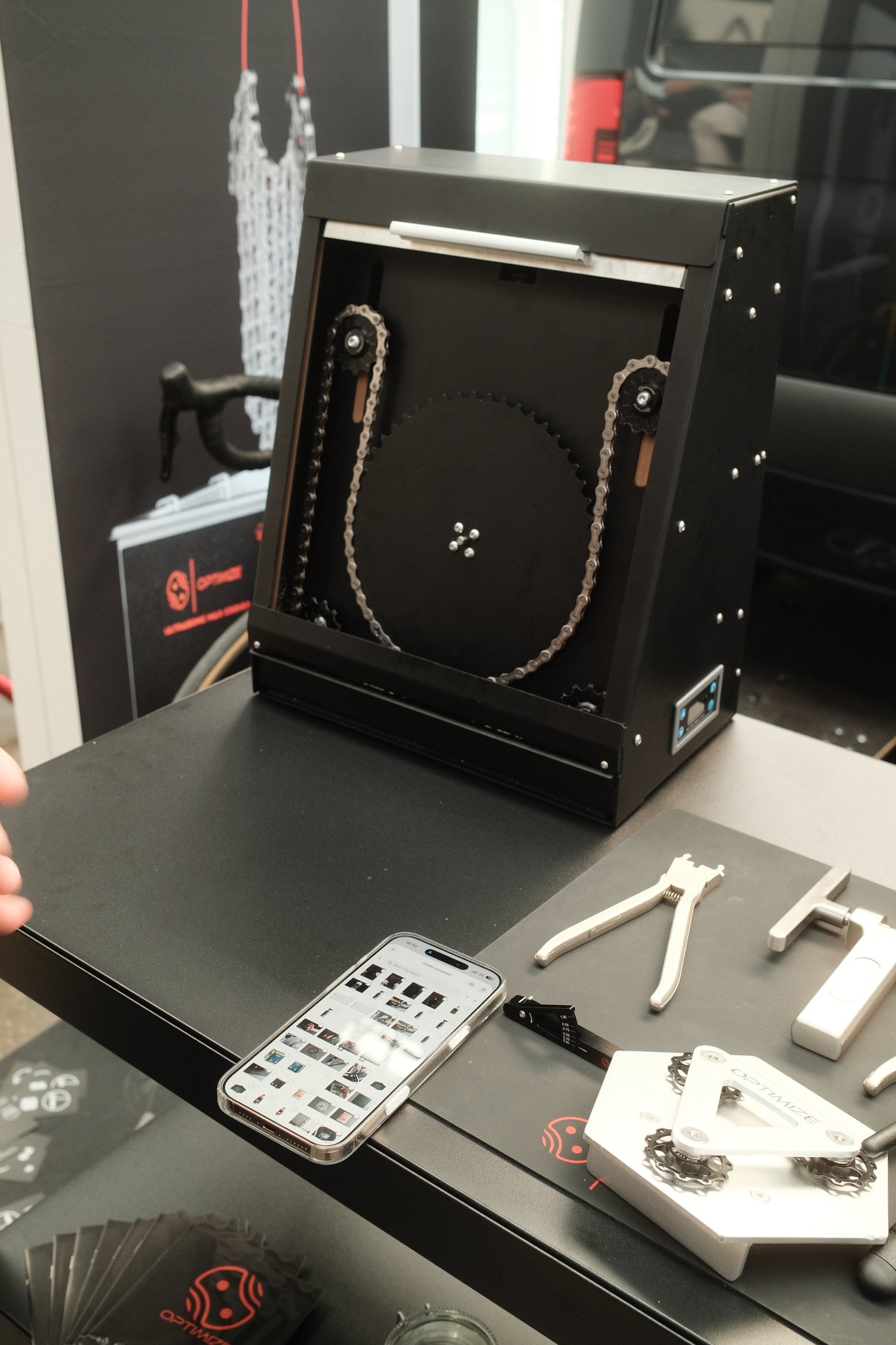 Der Optimize Kettenbrecher elektrisch von Optimize GmbH, ein schwarzes automatisches Gerät, reinigt Ihre Fahrradkette in seiner Box. Werkzeuge, Anleitungen und eine Telefongalerie liegen auf dem Tisch - alles, was du brauchst, um deine Kette renntauglich zu halten.