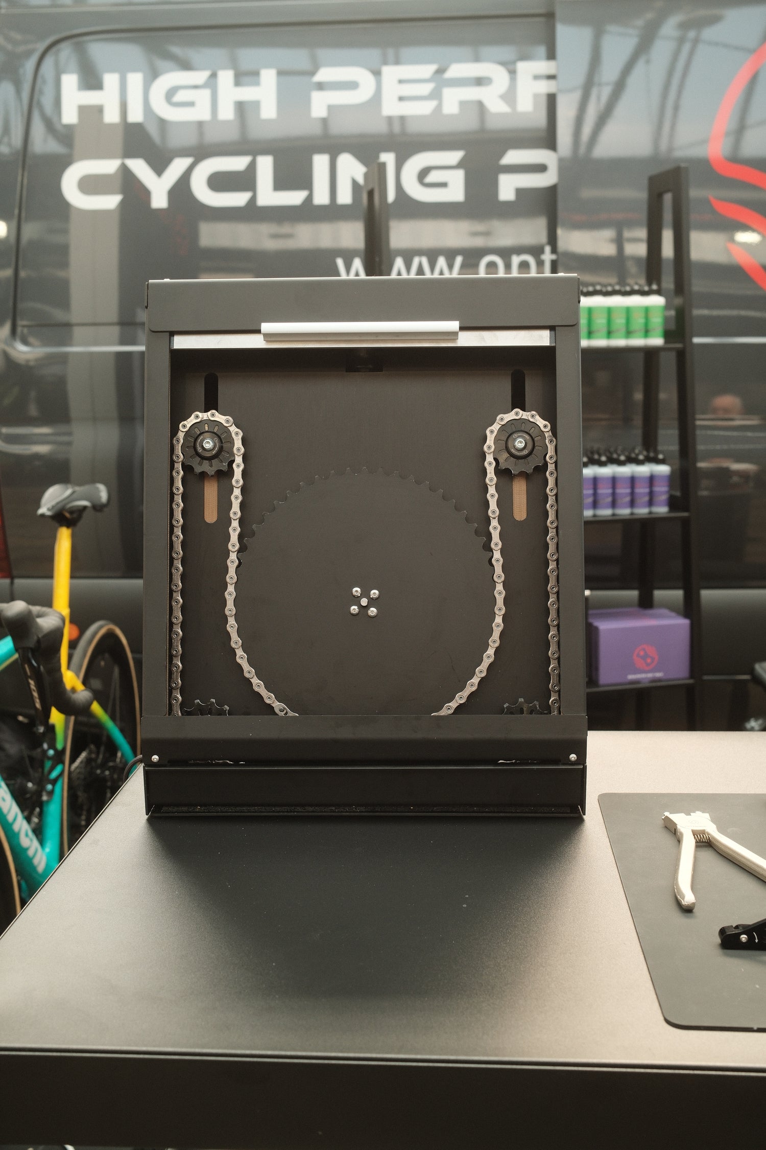Eine Nahaufnahme des Optimize GmbH Optimize Kettenbrecher elektrisch Radsport-Displays, mit einem großen Zahnrad und einer Kette auf beiden Seiten, Werkzeugen in der Nähe und einem High Perf Cycling-Banner, demonstriert die Kettenoptimierung für Spitzenleistungen im Rennen.