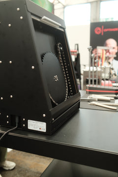 Der Optimize Kettenbrecher elektrisch der Optimize GmbH, eine schwarze Industriemaschine mit großem Sägeblatt und Kettenmechanismus mit elektrischem Ketteneinlaufsystem, wird auf einem Tisch in einer Ausstellung mit Werkzeugen und unscharfem Hintergrund präsentiert.