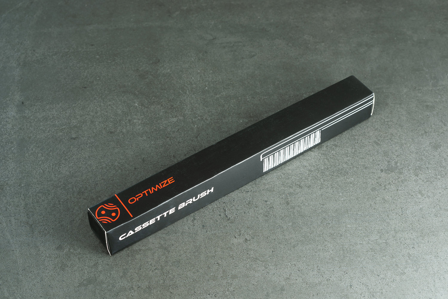 Eine lange, rechteckige schwarze Schachtel mit der Aufschrift Optimize Kassetten-Reinigungsbürste der Optimize GmbH, einem Barcode und einem roten Logo liegt auf einer grauen, strukturierten Fläche.