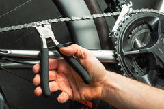Eine Hand benutzt die „Optimize Kettenschloss-Zange“ der Optimize GmbH, ein spezielles Fahrradketten-Werkzeug „Made in Germany“, um eine Fahrradkette im Bereich der Schaltung und der Kurbelgarnitur zu reparieren oder einzustellen.