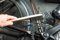 Mit der Optimize Kassetten-Reinigungsbürste der Optimize GmbH mit Holzgriff reinigt eine Hand gründlich Kette, Schaltung und Pedale eines dunkelfarbigen Fahrrads.
