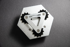 Der „Kettenbrecher mechanisch“ von Optimize GmbH ist ein metallisches, sechseckiges Objekt mit drei schwarzen Zahnrädern, einem dreieckigen Metallteil mit der Gravur „OPTIMIZE“ sowie Designelementen, die von einem mechanischen Kettenbrecher inspiriert sind, vor einem dunkelgrauen Hintergrund.