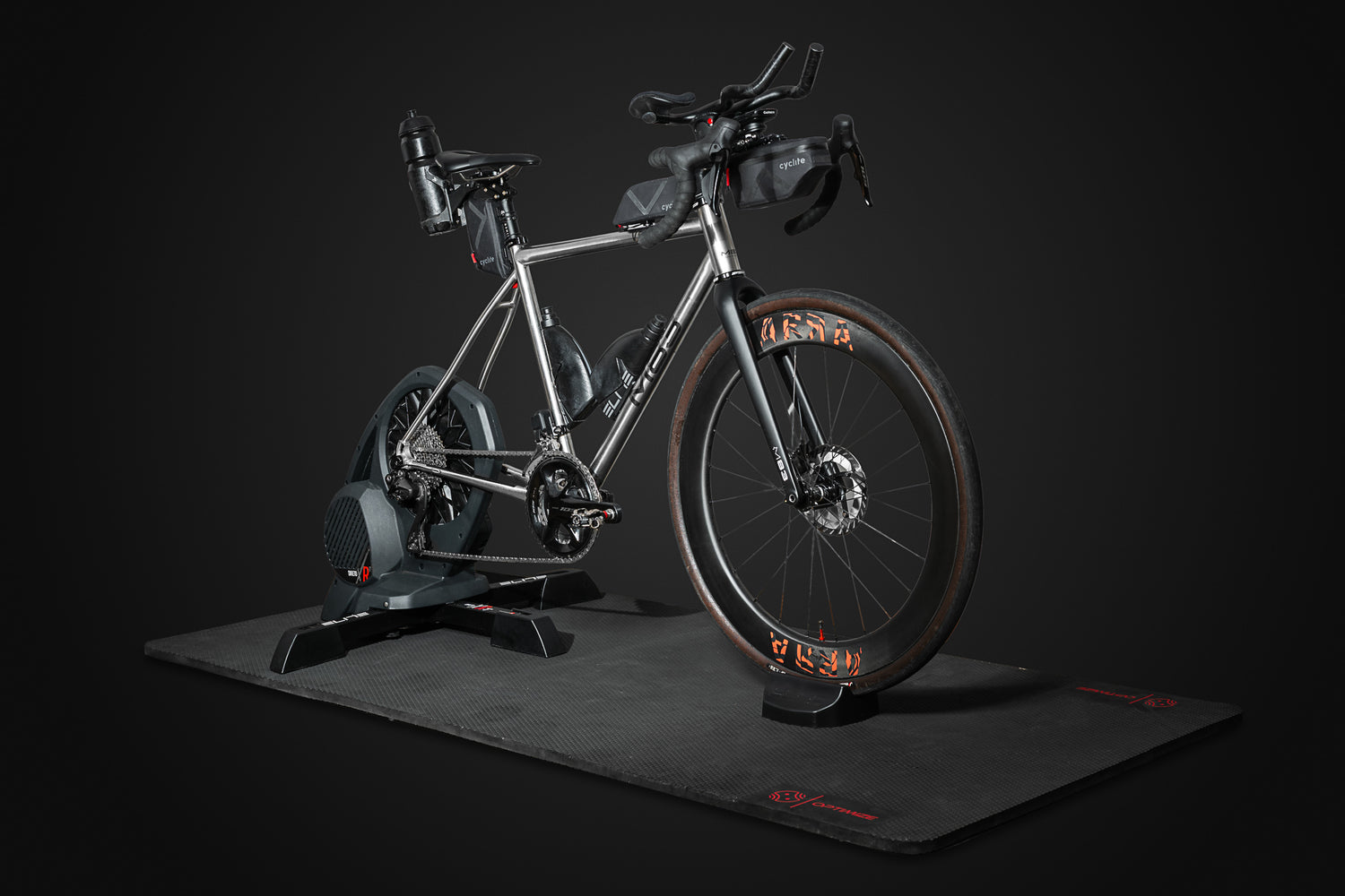 Eine „Indoor Matte“ der Optimize GmbH in eleganter, matter Optik mit roten Akzenten bildet die Kulisse für ein silbernes Rennrad, das auf einem Smart-Trainer montiert ist und vor einem dunklen Hintergrund steht. Am Rahmen und am Lenker des Fahrrads sind verschiedene Zubehörteile angebracht.