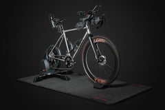 Eine „Indoor Matte“ der Optimize GmbH in eleganter, matter Optik mit roten Akzenten bildet die Kulisse für ein silbernes Rennrad, das auf einem Smart-Trainer montiert ist und vor einem dunklen Hintergrund steht. Am Rahmen und am Lenker des Fahrrads sind verschiedene Zubehörteile angebracht.