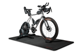Ein silbernes Rennrad, das auf einem Smart-Trainer montiert ist, steht auf der „Optimize GmbH Indoor Matte“, die sich durch ein elegantes schwarzes Design auszeichnet. Das Fahrrad ist mit Trinkflaschen, Lenkertaschen und auffälligen orangefarbenen Reifenakzenten ausgestattet. Der Hintergrund ist schlicht weiß.