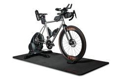Ein silbernes Rennrad, das auf einem Smart-Trainer montiert ist, steht auf der „Optimize GmbH Indoor Matte“, die sich durch ein elegantes schwarzes Design auszeichnet. Das Fahrrad ist mit Trinkflaschen, Lenkertaschen und auffälligen orangefarbenen Reifenakzenten ausgestattet. Der Hintergrund ist schlicht weiß.