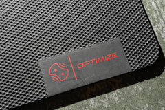 Eine Nahaufnahme eines strukturierten schwarzen „Indoor Matte“-Produkts der Optimize GmbH, auf dem ein Etikett mit einem roten Logo und dem Schriftzug „OPTIMIZE“ in Rot auf einem grünen Untergrund zu sehen ist.