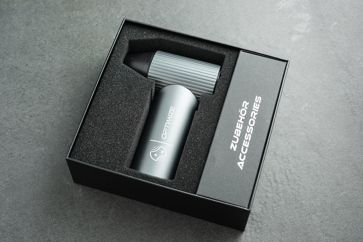 Ein elegantes silbernes Gerät mit der Aufschrift „Optimize GmbH“ liegt ordentlich in einer schwarzen Schaumstoffverpackung neben einem gerippten Metallteil – einer wesentlichen Komponente des „Optimize Akku Luftgebläse“ – und einer Schachtel mit der Aufschrift „ZUBEHÖR ACCESSORIES“ auf einer grauen Unterlage.