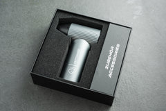 Ein elegantes silbernes Gerät mit der Aufschrift „Optimize GmbH“ liegt ordentlich in einer schwarzen Schaumstoffverpackung neben einem gerippten Metallteil – einer wesentlichen Komponente des „Optimize Akku Luftgebläse“ – und einer Schachtel mit der Aufschrift „ZUBEHÖR ACCESSORIES“ auf einer grauen Unterlage.